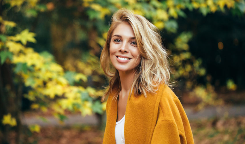 Blonde woman smiles free of periodontal disease while outside in the fall foliage