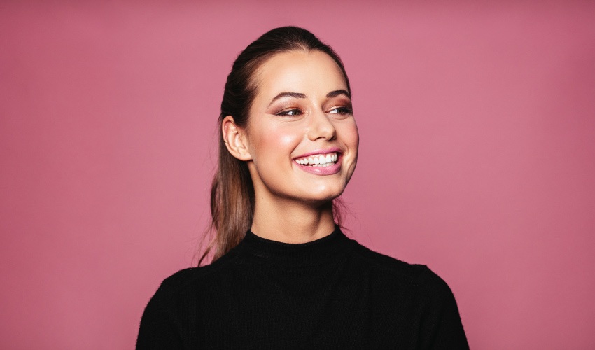 Brunette woman in a black shirt smiles against a pink background after whitening her teeth